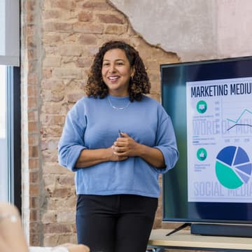 Woman stands in boardroom presenting marketing mediums on the projector screen.